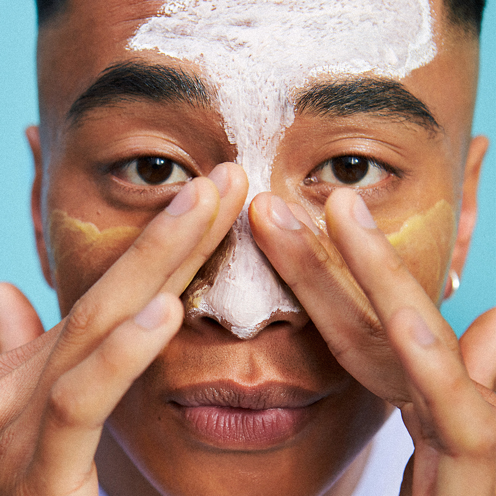 Närbild på en person som applicerar ansiktsmask i olika färger på pannan, näsan och kinderna.